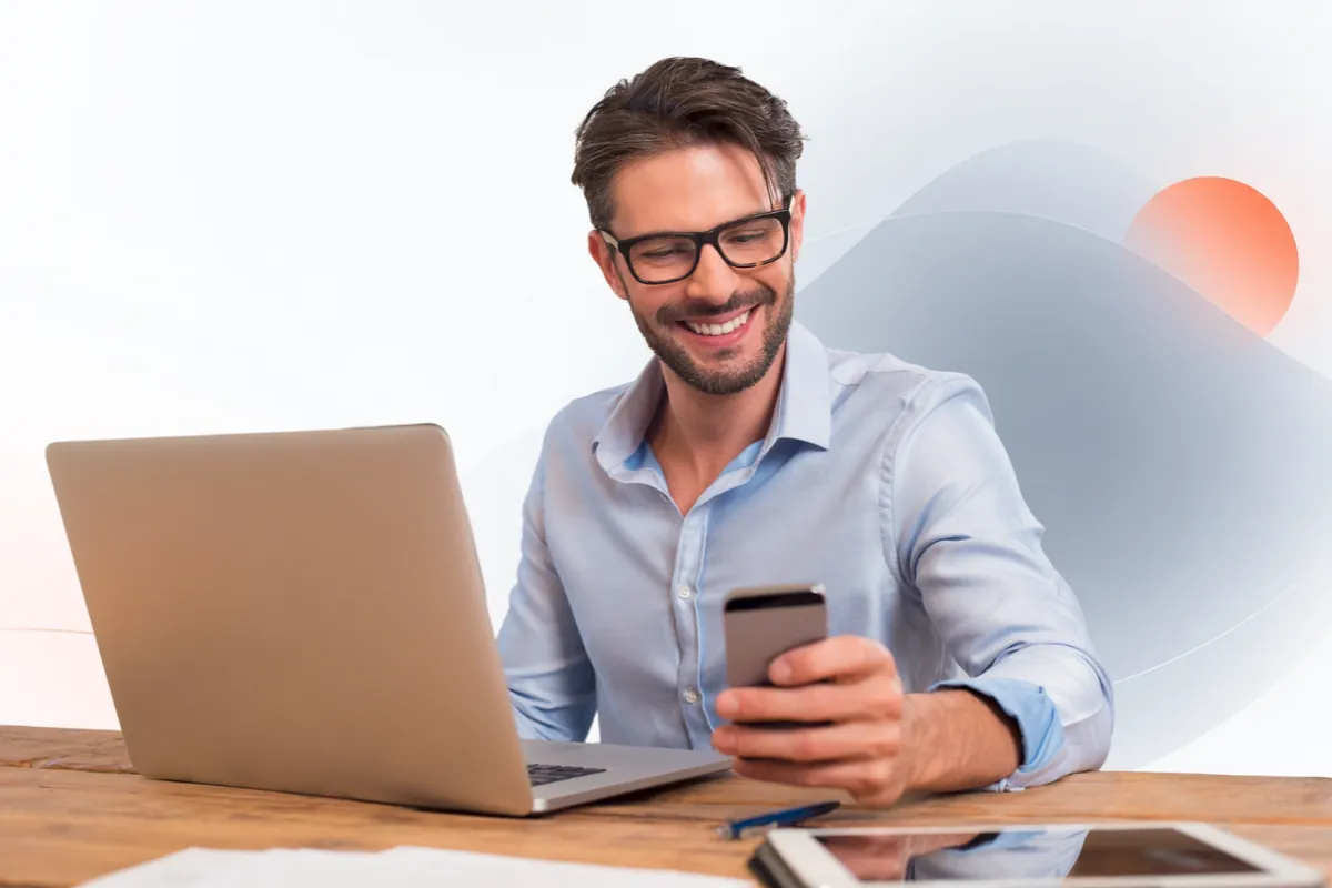 Lächelnder Mann am Laptop schaut auf sein Smartphone